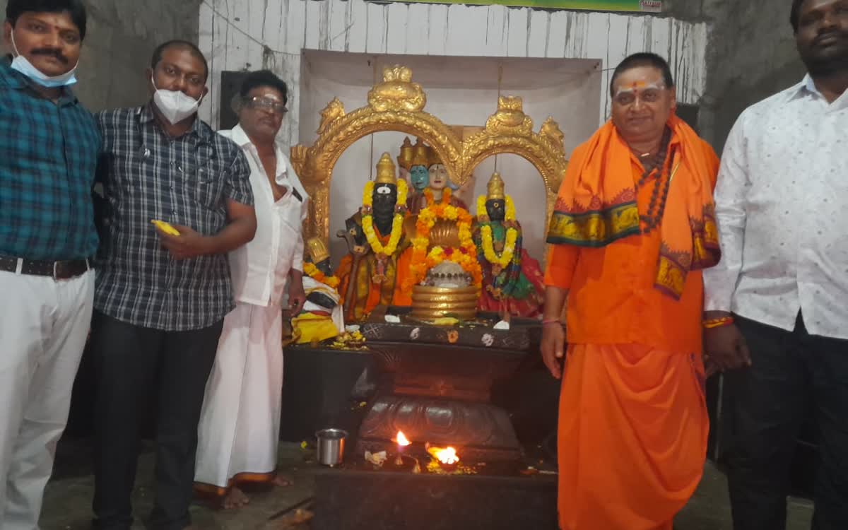 Guruji standing at the shrine with devotees, the lamps lit before the deities